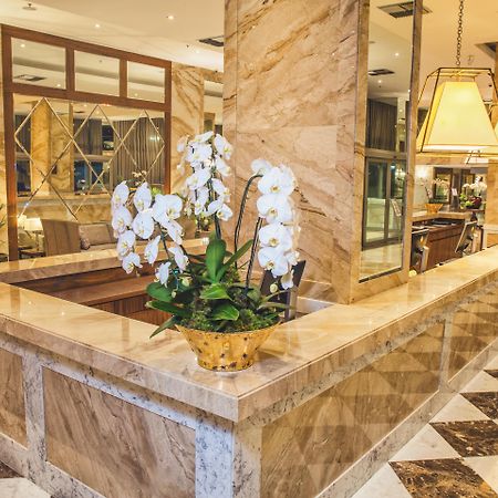 Windsor Marapendi Hotel Ρίο ντε Τζανέιρο Εξωτερικό φωτογραφία The photo depicts a luxurious hotel lobby featuring a polished marble reception desk. There are elegant white orchids arranged in a gold pot on the counter, adding a touch of sophistication. The surrounding interior showcases marble walls, stylish li