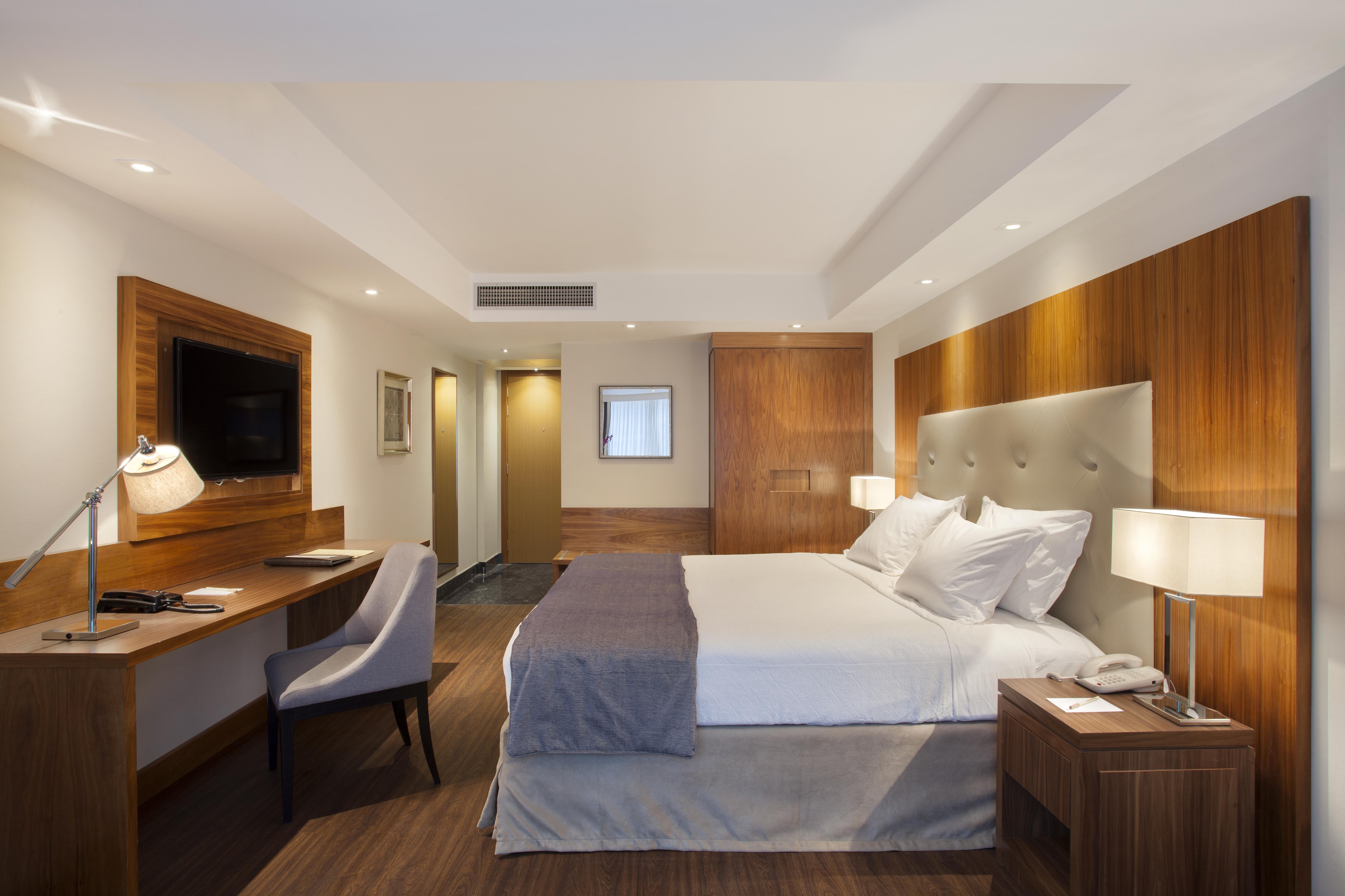 Windsor Marapendi Hotel Ρίο ντε Τζανέιρο Εξωτερικό φωτογραφία The photo shows a modern hotel room featuring a large bed with white and gray bedding. The headboard is upholstered in light fabric. There are two bedside lamps on either side of the bed, providing warm lighting. A television is mounted on the wall o